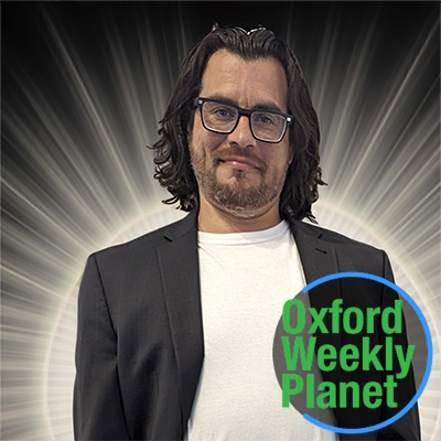 Smirking man with dark hair and glasses with the Oxford Weekly Planet logo in the foreground