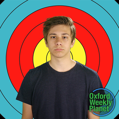 Glum teen boy with medium brown hair in front of an archery target with the Oxford Weekly Planet logo in the foreground