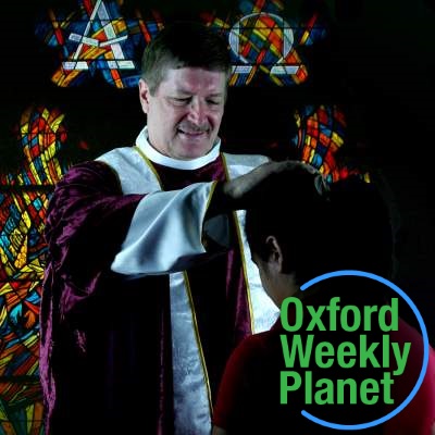 Man in minister's robes blessing a kneeling woman with a stained glass window in the background with the Oxford Weekly Planet logo in the foreground