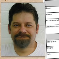 Man with dark hair, mustache, and goatee and a wry smile with a criminal history form in the background