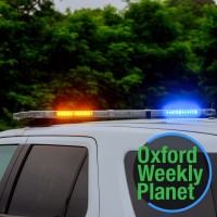 Police patrol vehicle with lights on and trees in the background and the Oxford Weekly Planet logo in the foreground