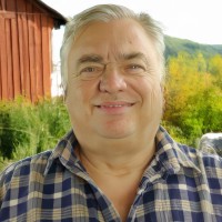 Large silver-haired man wearing a plaid shirt and standing in front of a rusted antique vehicle