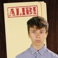 Serious teen boy with dark hair in front of a manila folder stamped 'Alibi'