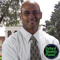 Smiling bald man wearing glasses with the Oxford Weekly Planet logo in the foregground