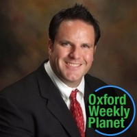 Husky smiling man in a suit with the Oxford Weekly Planet logo in the foreground