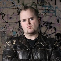 Sullen young man with short dark hair and facial hair, wearing a black leather jacket