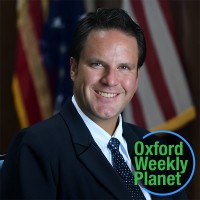 Husky smiling man in a suit with the Oxford Weekly Planet logo in the foreground