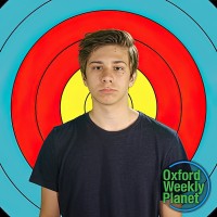 Glum teen boy with medium brown hair in front of an archery target with the Oxford Weekly Planet logo in the foreground