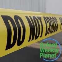 Close-up of crime scene tape at a scene with the Oxford Weekly Planet logo in the foreground