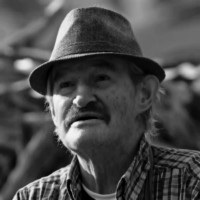 Older man with gray hair and a dark mustache, wearing a fedora