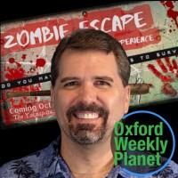 Smiling man with a goatee in front of a Zombie Apocalypse poster with the Oxford Weekly Planet logo in the foreground