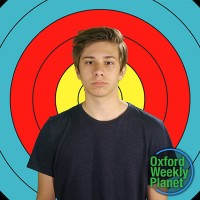 Glum teen boy with medium brown hair in front of an archery target with the Oxford Weekly Planet logo in the foreground