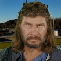 Serious man with long dark hair and facial hair, wearing a detective windbreaker