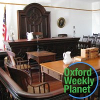 Sunlit courtroom with the Oxford Weekly Planet logo in the foreground