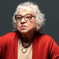 White-haired woman with dark-rimmed glasses, looking displeased