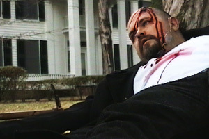 Man with a bloody head wound leaning against a tree with Rowan Oak in the background