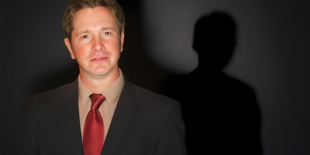 Serious man with short brown hair with the silhouette of an unidentified man behind him