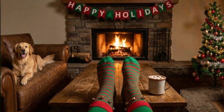 Feet in socks, resting on a table in front of a fire in a fireplace. There's a Happy Holidays banner above the fireplace and a golden retriever lying on a nearby sofa