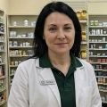 Slightly smiling woman with black hair, wearing a lab coat and standing in a pharmacy