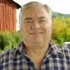 Large silver-haired man wearing a plaid shirt and standing in front of a rusted antique vehicle