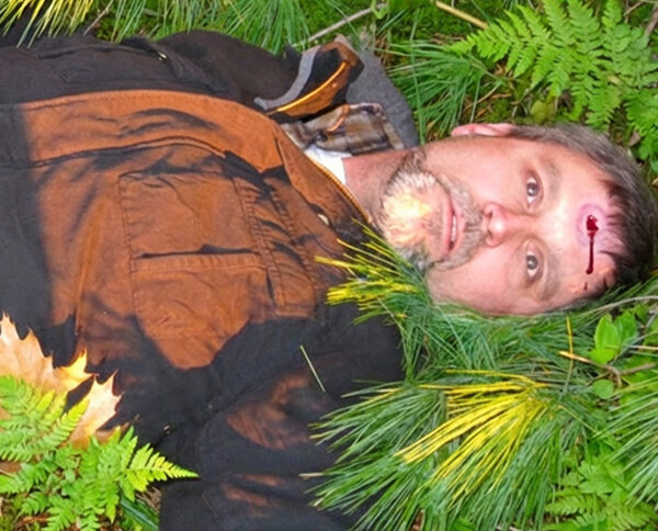 Man with a bullet wound in his forehead, lying on the ground surrounded by pine needles and ferns