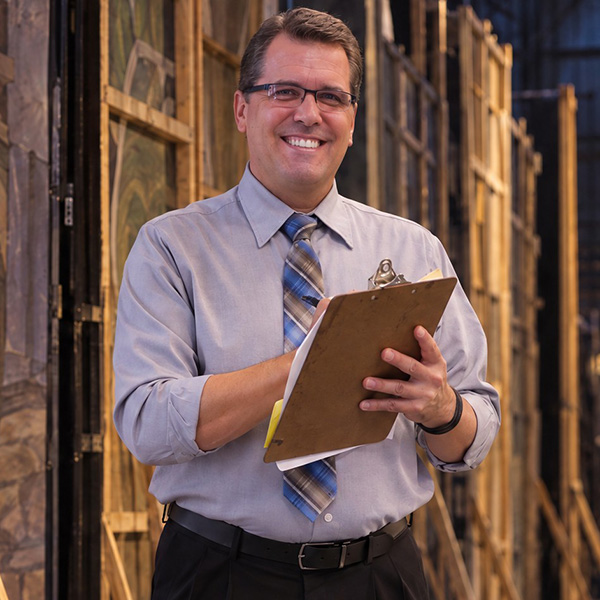 Smiling, clean-cut man with glasses, holding a clipboard and standing backstage