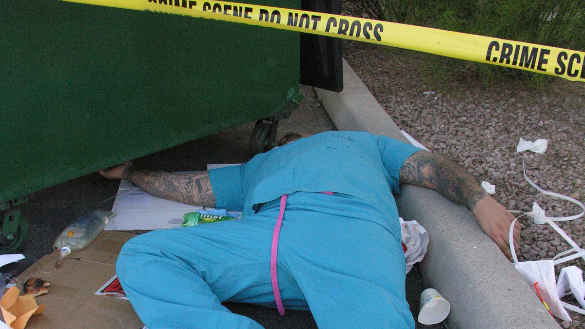 Man in hospital scrubs lays dead by a green dumpster