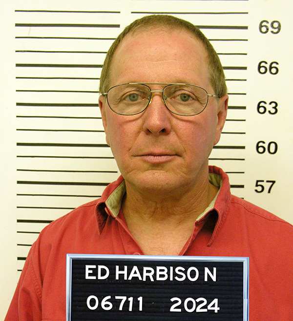 Man with dark blond receding hair and glasses holding a letterboard with the name "Ed Harbison" and standing in front of a height chart