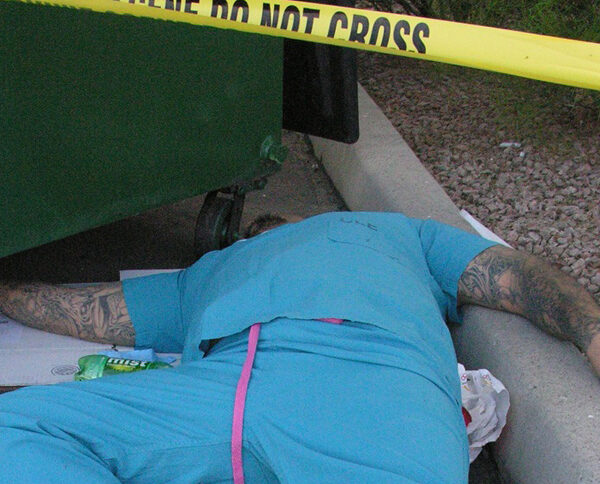 Man in hospital scrubs lays dead by a green dumpster