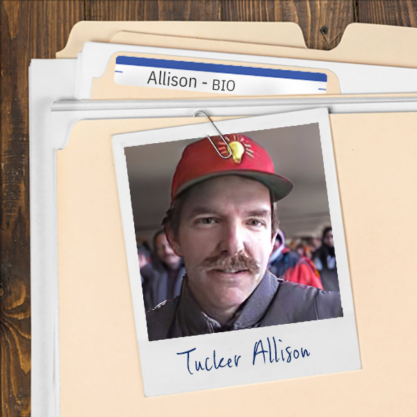 We see a file folder on a desk. Clipped to the folder is a picture of a man with brown hair and mustache wearing a red baseball cap