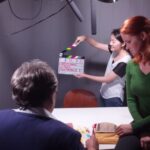 A picture of a man and woman sitting near a table, with a woman in the background holding an open clapboard