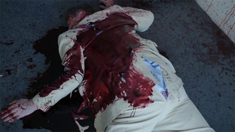 Dead man wearing beige suit covered in blood laying in prone position. Blood surrounds body. Rip in back of mans pants exposes underwear.