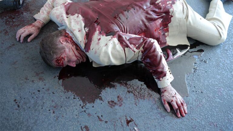 Dead man wearing beige suit covered in blood laying in prone position. Blood surrounds body.