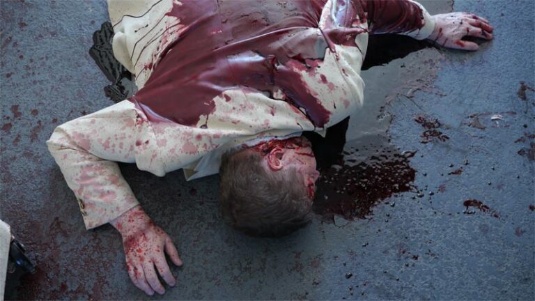 Dead man wearing beige suit covered in blood laying in prone position. Blood surrounds body.