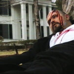 Man with a bloody head wound leaning against a tree with Rowan Oak in the background