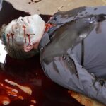 A picture of a pale body with blood pooling around him.