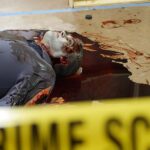 A picture of a pale body behind a crime scene tape with blood pooling around him.