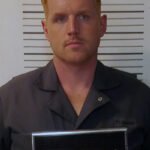 a man wearing a grey button down polo shirt with blond spiked hair standing in front of a height chart and holding up a card for a mugshot. Name appears as "Emmett Sanford"