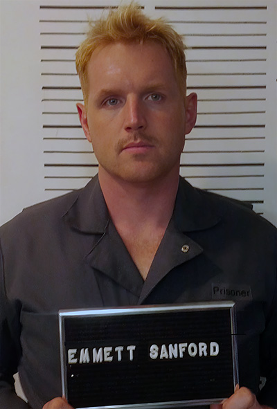 a man wearing a grey button down polo shirt with blond spiked hair standing in front of a height chart and holding up a card for a mugshot. Name appears as "Emmett Sanford"
