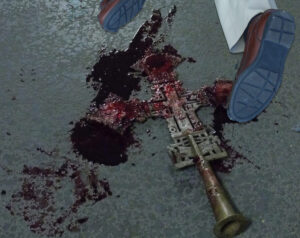 legs of person wearing beige pants and brown shoes laid in a prone position. Blood under and around both feet. Bloody gold colored metal cross surrounded by blood under feet.