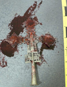 Gold tone metal cross surrounded by blood on floor. Yellow evidence ruler partially exposed on the right side of cross.