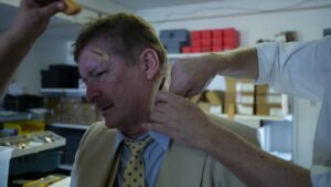 Male with mustache wearing a yellow suit and yellow polka dot tie having fake neck wound being applied by person wearing white coat. The sleeves of the white coat are folded midway up arm.