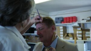 Male with mustache wearing a yellow suit having a fake wound applied on left temple by lady with grey hair and glasses. The lady is holding a paintbrush to apply the fake wound.