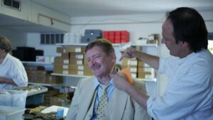 smiling male with mustache wearing a yellow suit with a yellow polka dot tie. Fake wound being applied on neck of male in yellow suit by a male with black hair wearing a white coat. Boxes in the background.