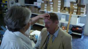 male with yellow suit and yellow polka dot tie having fake wound applied to left temple by lady with grey hair and glasses. Empty boxes in the background behind the male.