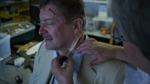Male in a yellow suite with wound on left temple. Lady with grey hair and glasses applying fake wound to the neck area of the male.