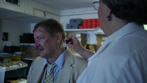 Male with mustache in a yellow suite with a yellow polka dot tie. Wound on left temple and straight wound from chin to ear being applied by lady in a white coat with glasses.