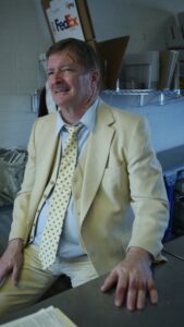 Male with mustache in a yellow suit with a yellow polka dot tie. The male's hand is laid flat on a desk and he has a smile.