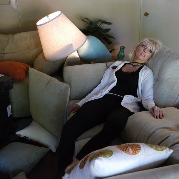 Blonde woman seated on sofa, eyes open and an electrical cord around her neck, with an overturned lamp in the background