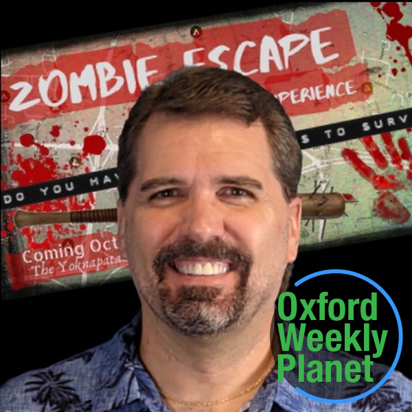 Smiling man with a goatee in front of a Zombie Escape poster with the Oxford Weekly Planet logo in the foreground